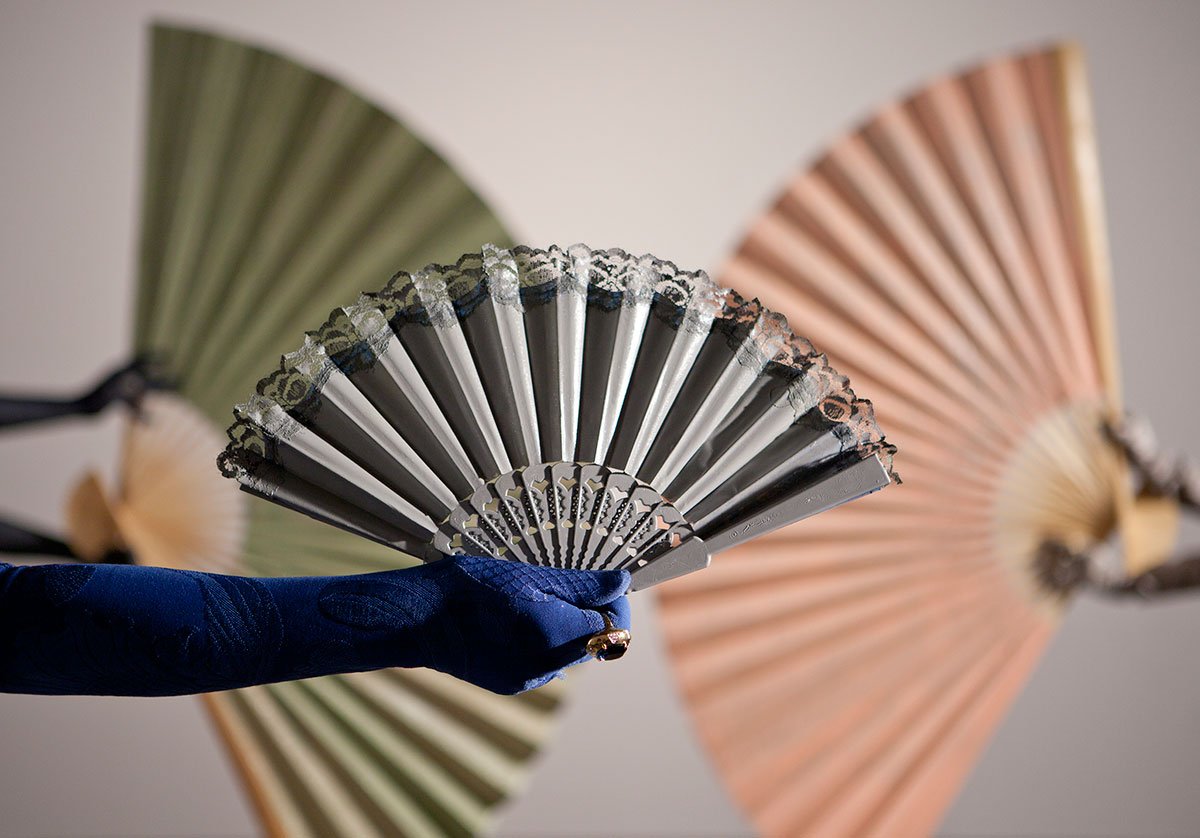 Arm with blue glove holding lace fan against background of green and pink fans