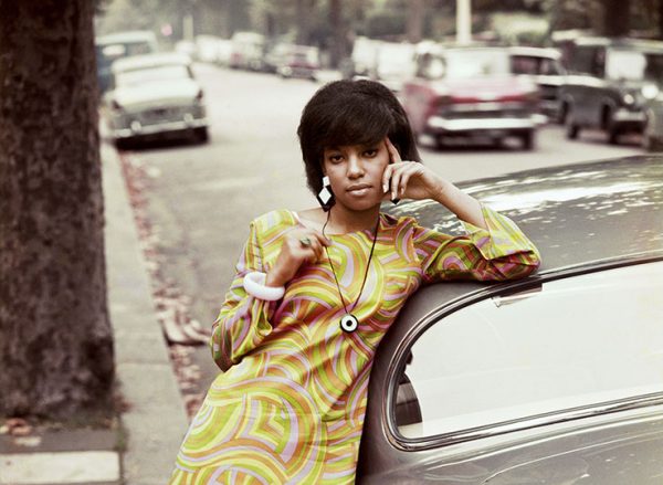 A woman waring a yellow dress leans glamourously on a silver car